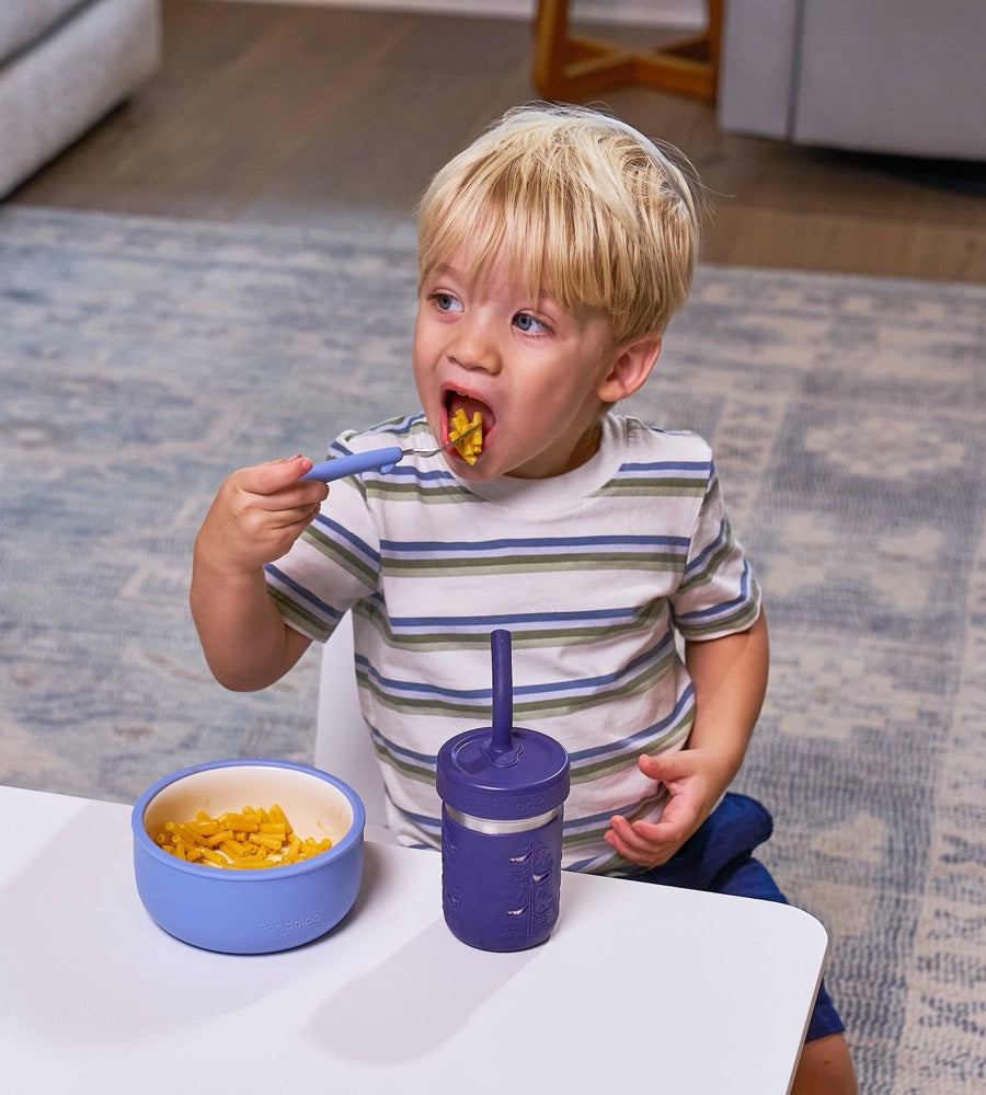 Toddler eating pasta with a Pandaloo Kids fork with a soft blue silicone handle, next to a Pandaloo porcelain bowl and kids cup